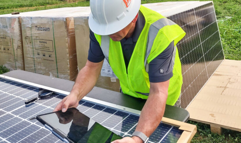 Solar energy technician wearing vest and helmet inspects solar panel using tablet outdoors.